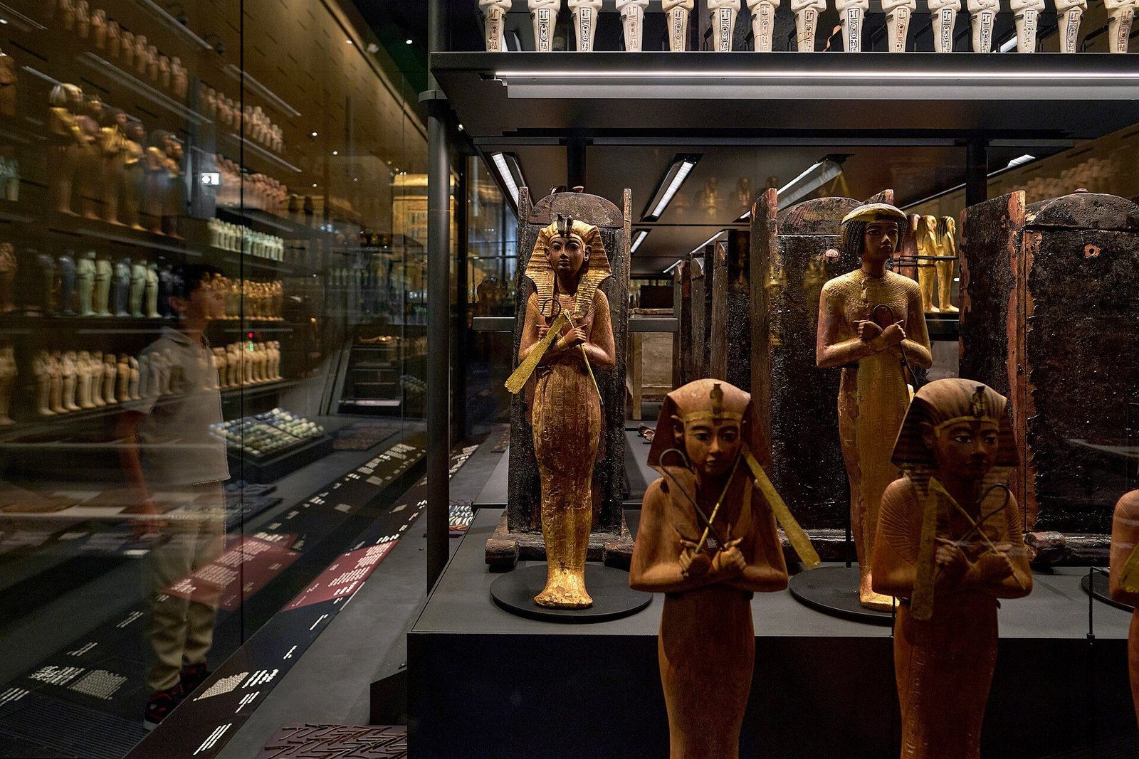 Estatuillas doradas y figuras funerarias de Tutankamón en las vitrinas de su galería del Gran Museo Egipcio, con un visitante reflejado en el cristal