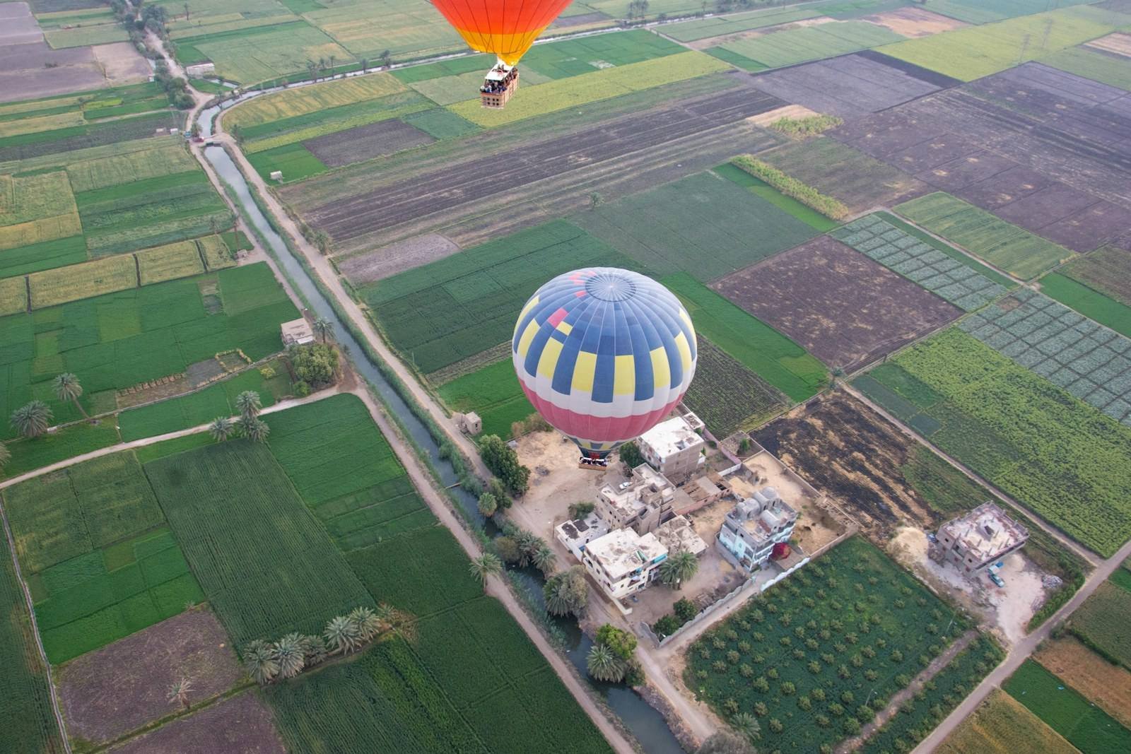 A hot air balloon floats above a patchwork of green and brown farm fields, a narrow canal and small flat roofed houses, with another balloon above