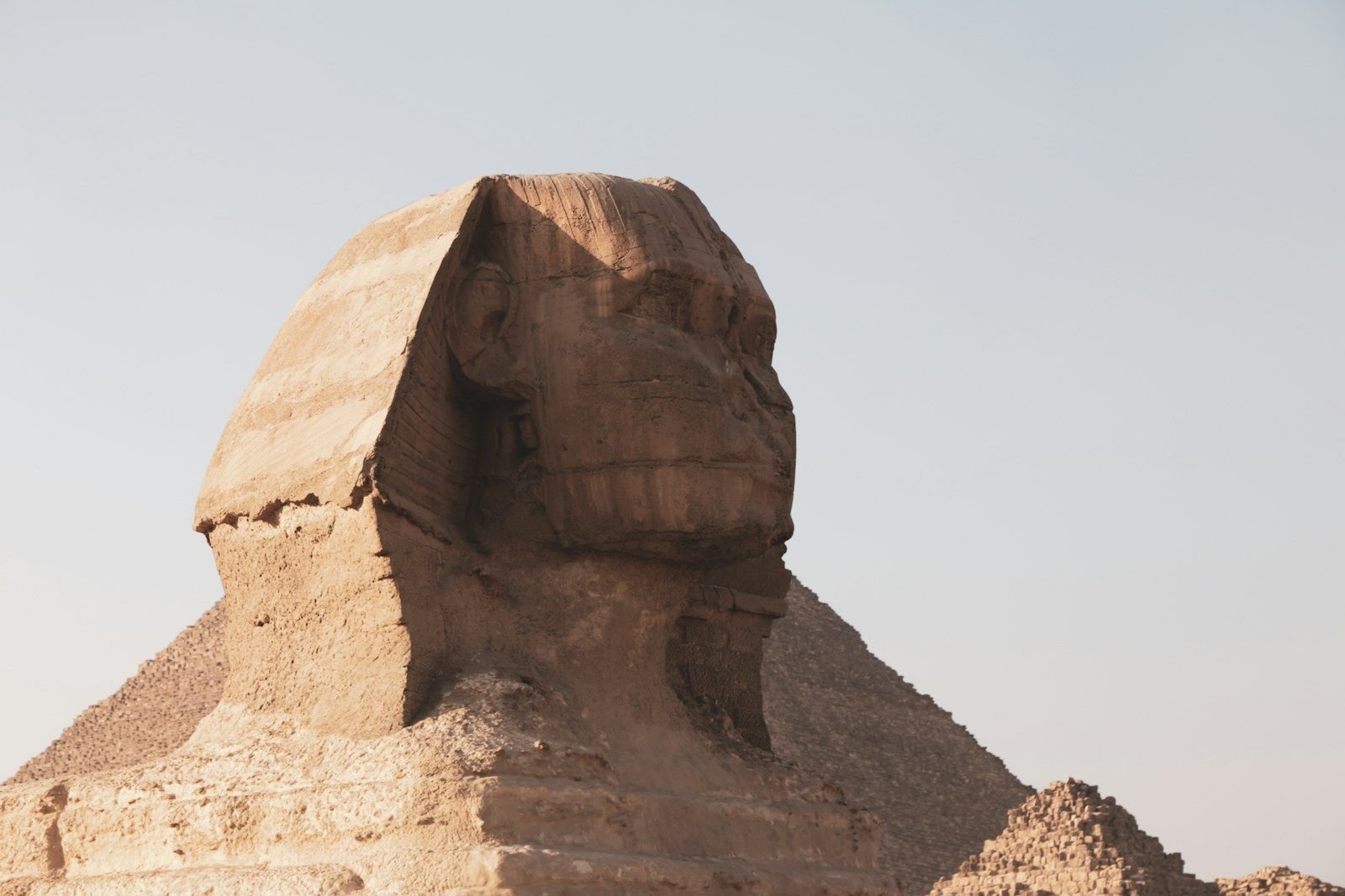 Primer plano de la cabeza de la Gran Esfinge de Giza, con el tocado a rayas y el rostro desgastado, y una pirámide detrás bajo un cielo claro