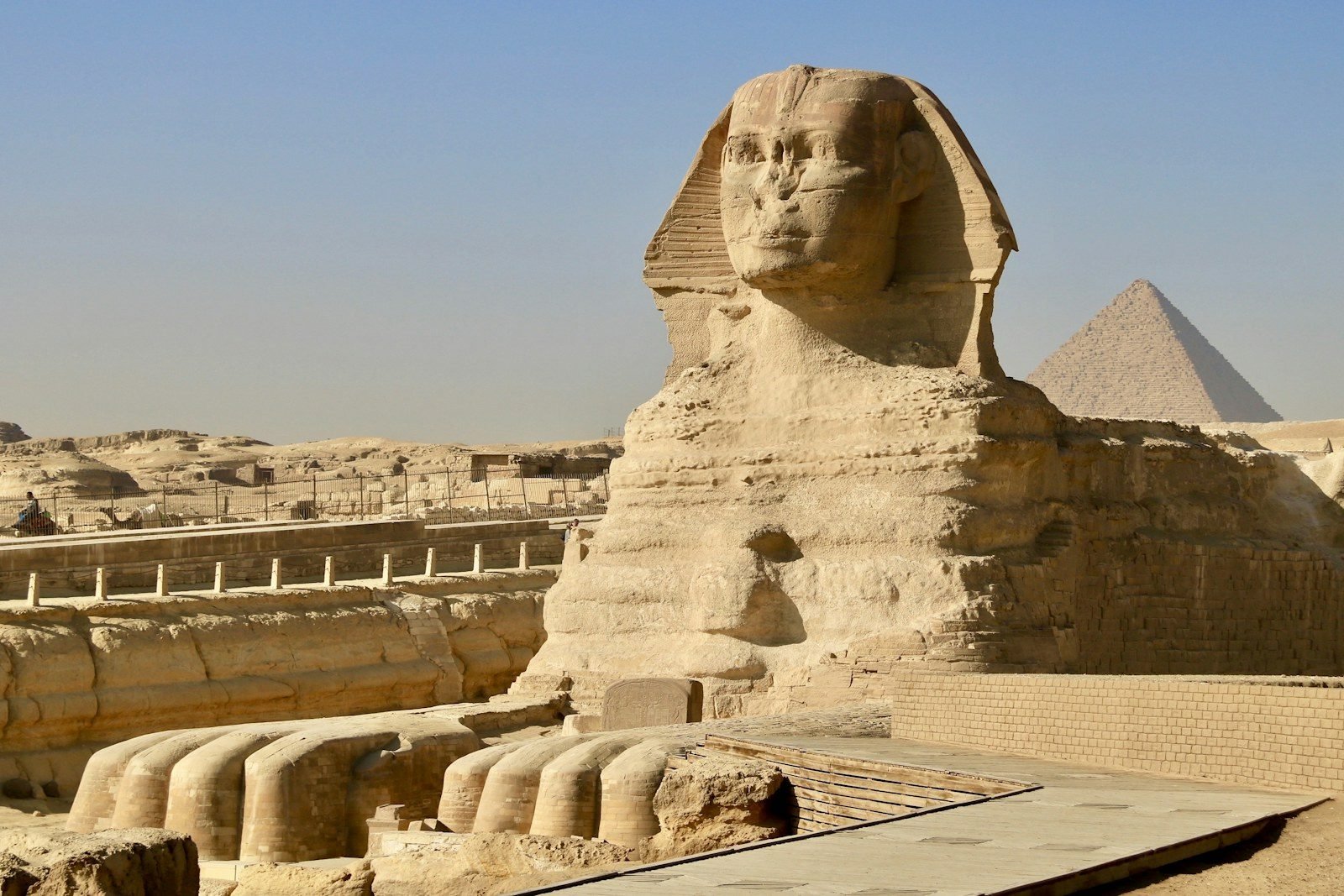 The Great Sphinx of Giza seen from the front, with its two forepaws, the enclosure walls and a wooden walkway, and a pyramid behind to the right under a clear blue sky.