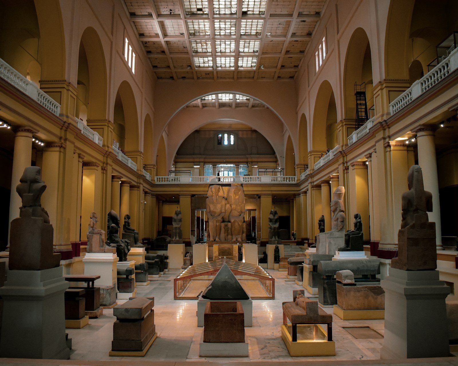 Amplia sala de museo con columnas ocres, balcones con balaustradas blancas y techo de casetones con claraboyas, llena de estatuas colosales, sarcófagos y una pirámide de piedra negra sobre un suelo de mármol.