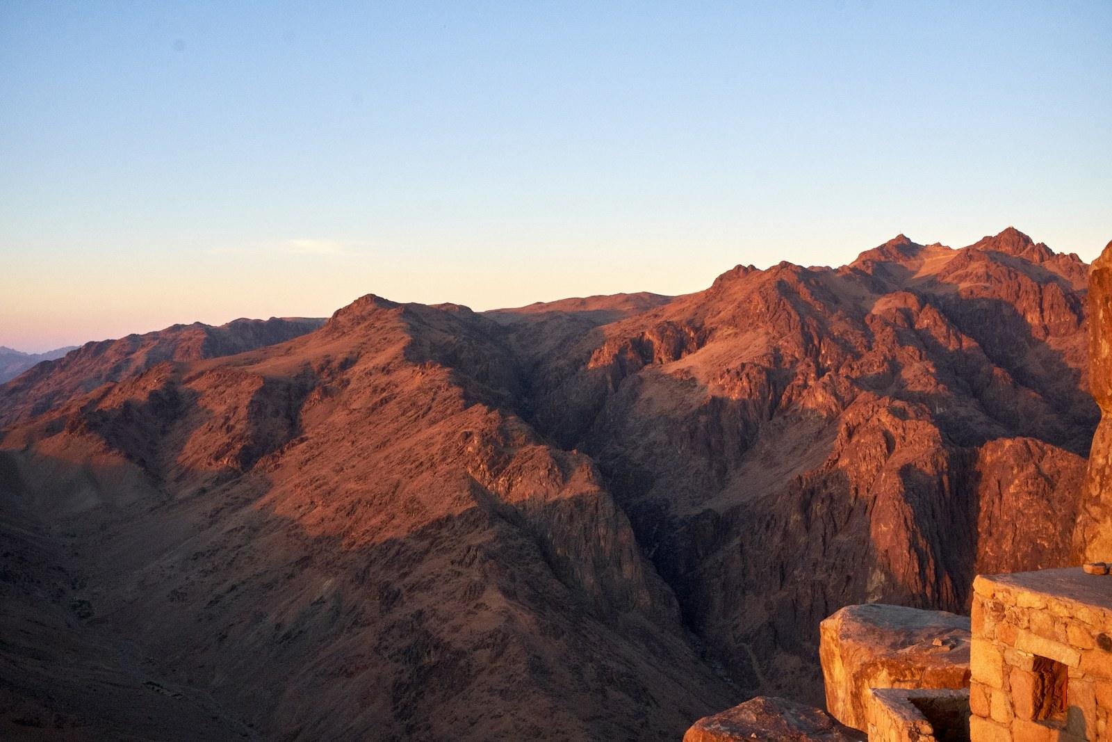 Laderas empinadas de montaña en tonos rojizos iluminadas por una luz cálida y baja, con un cielo pálido y un muro de piedra en la esquina