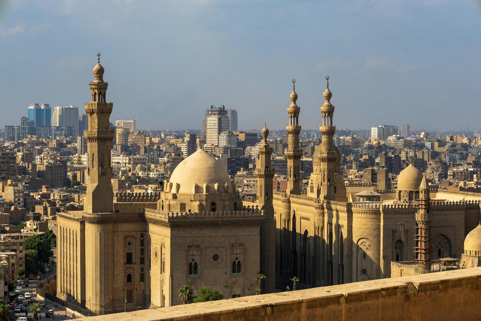 Vista elevada de dos grandes mezquitas con cúpulas y minaretes esbeltos entre los edificios de El Cairo, bajo un cielo azul grisáceo y luz cálida