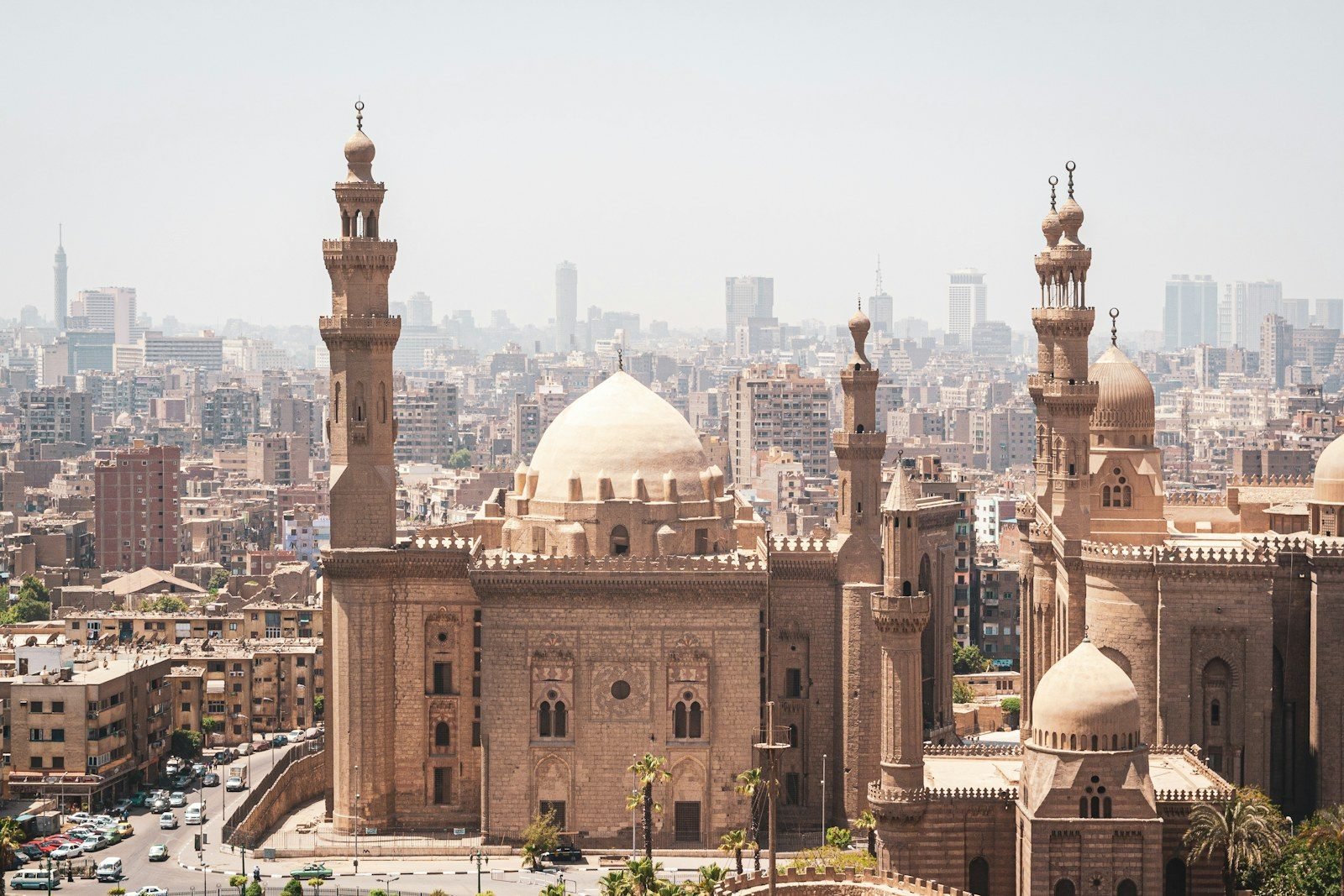 Vista elevada de dos grandes complejos de mezquitas de época mameluca, con cúpulas de piedra y minaretes esbeltos, sobre los tejados de El Cairo y una ligera bruma.