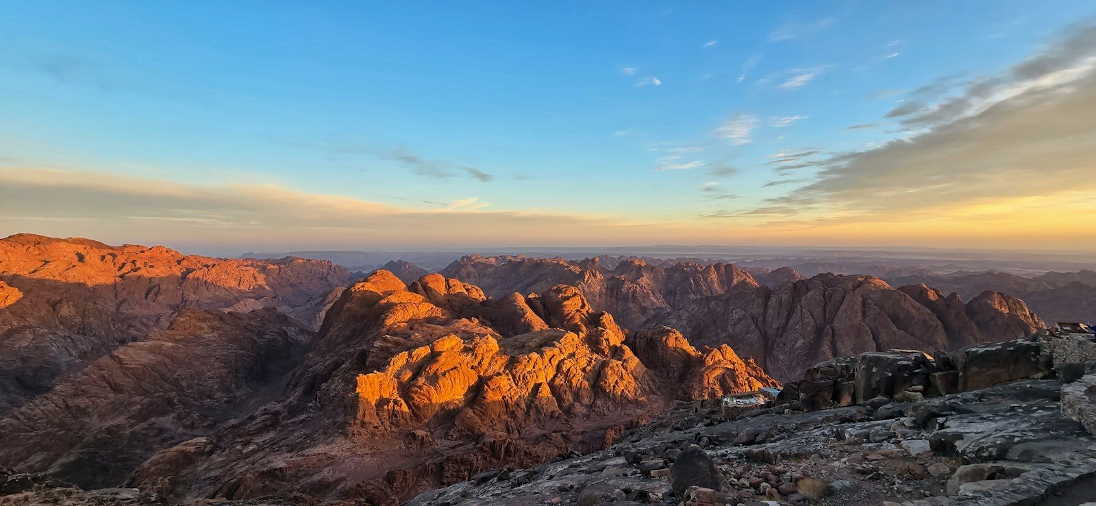 Panorama de montañas de granito rojizo iluminadas por una luz dorada desde una cima rocosa, con el cielo azul y franjas de nubes sobre el horizonte.