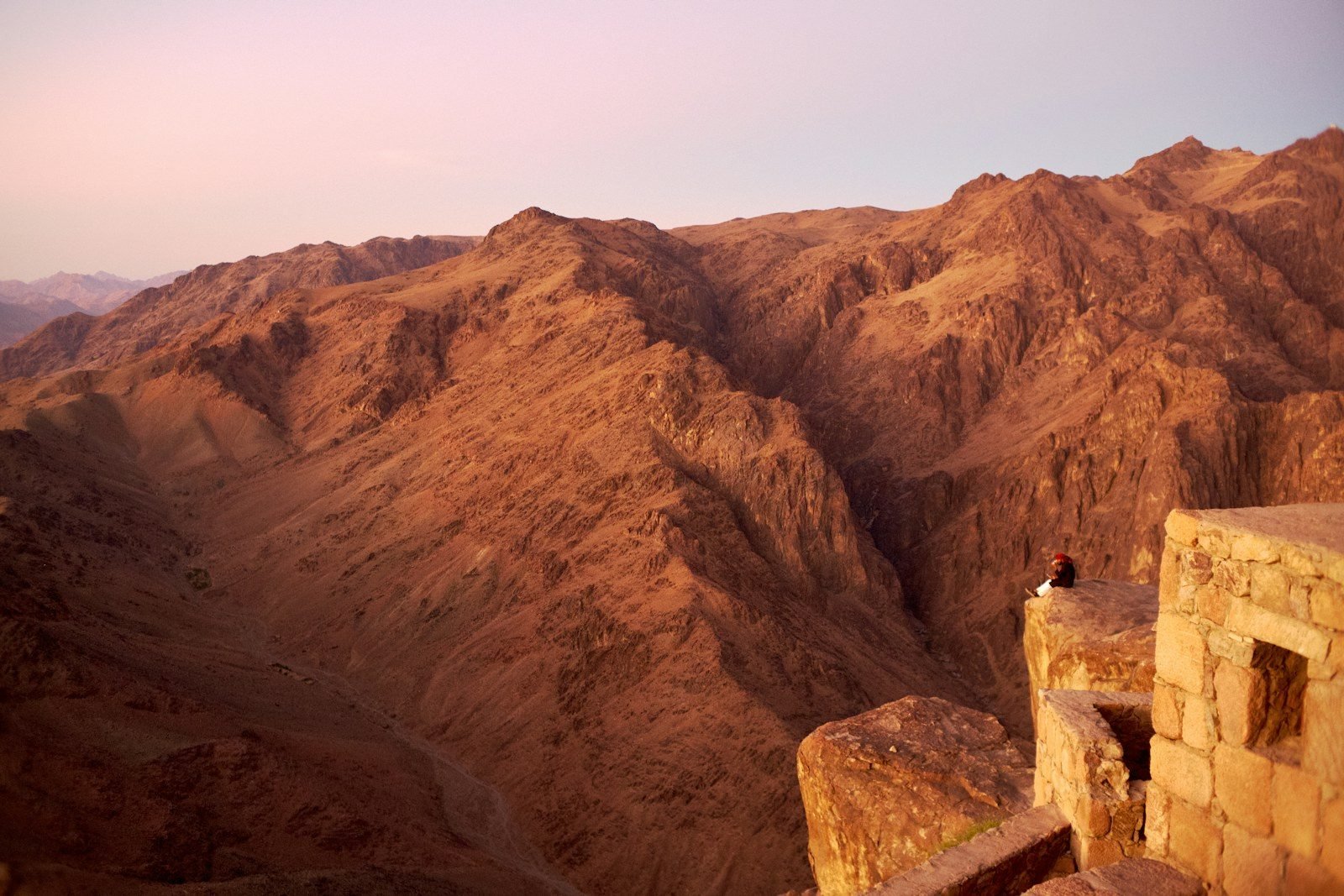 Montañas de roca rojiza y un valle profundo bajo un cielo rosado, vistos desde una terraza de piedra donde se sienta una figura diminuta.