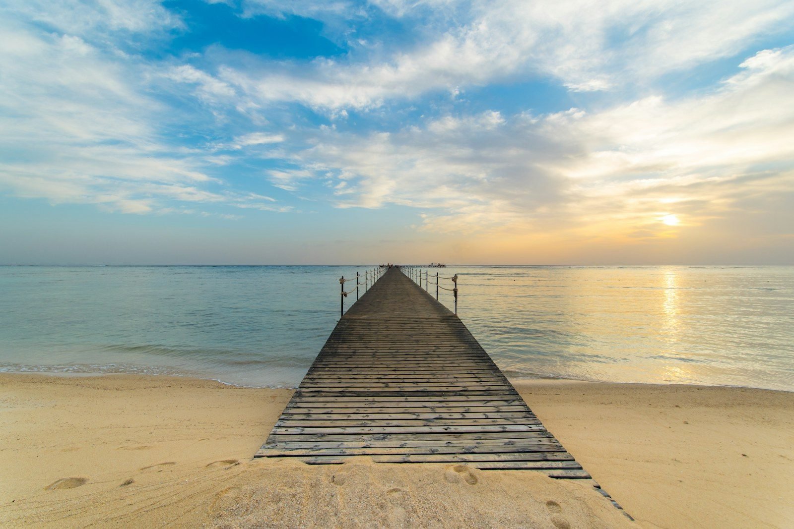 Un largo muelle de madera se adentra en un mar tranquilo desde una playa de arena fina, con el sol bajo entre las nubes y un reflejo dorado en el agua.