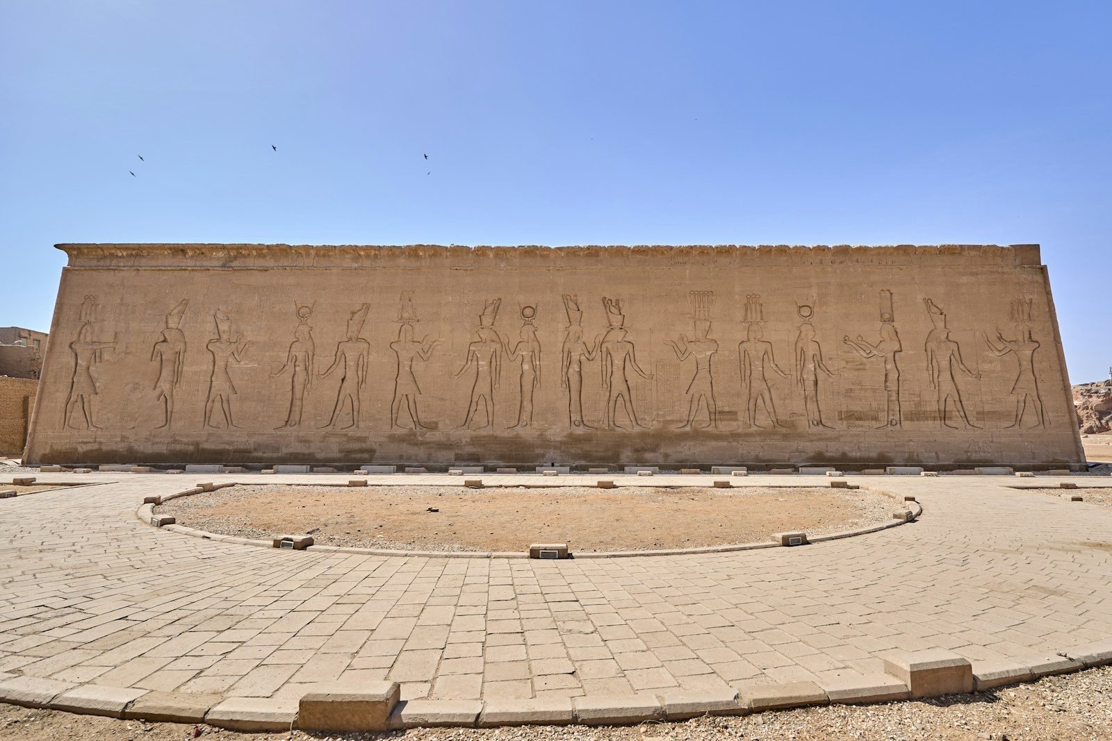 A long outer sandstone temple wall carved with a row of standing figures in sunk relief, under a clear blue sky.