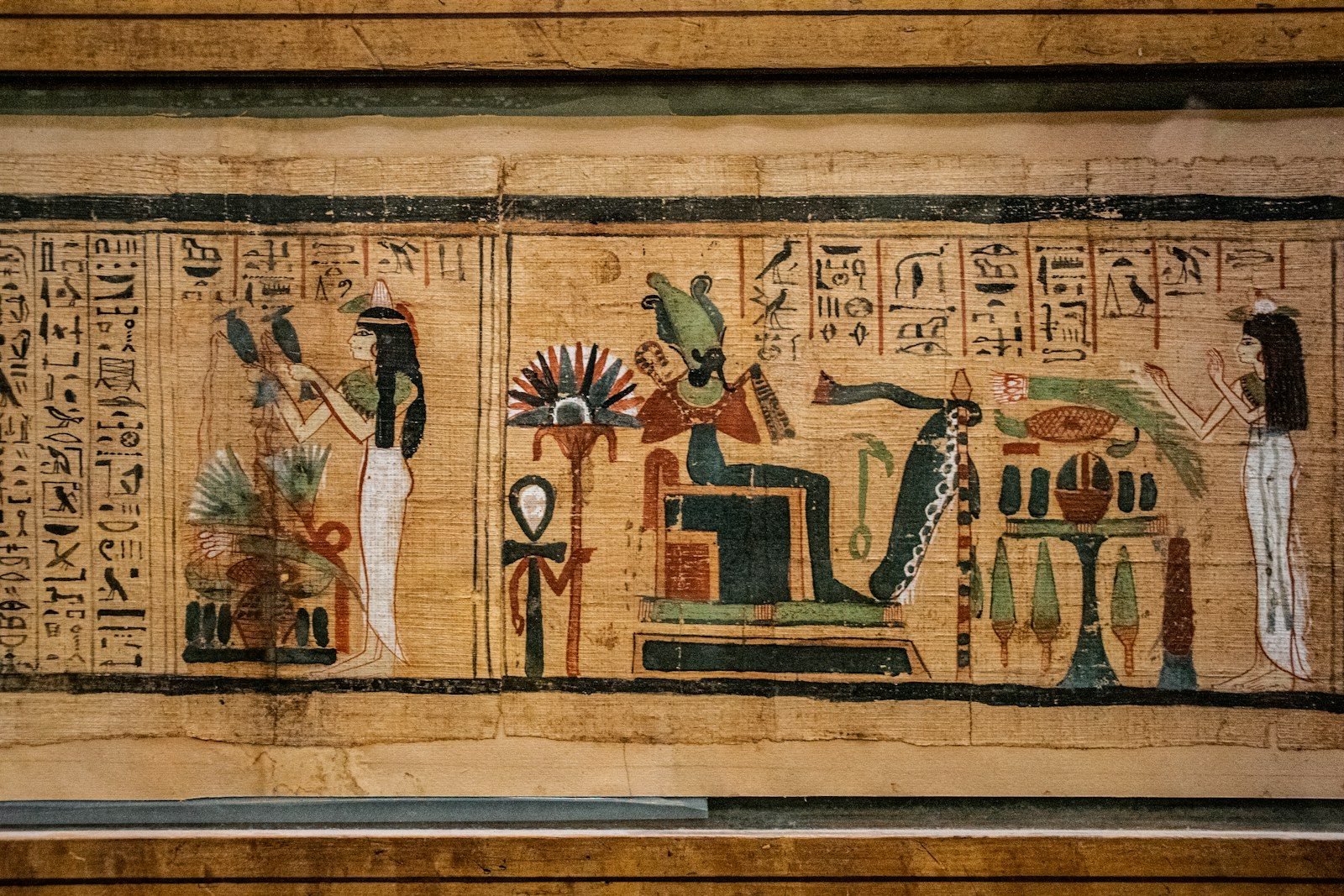 Papiro pintado tras un cristal, con un dios sentado de rostro verde y corona alta, dos figuras femeninas con ofrendas y columnas de jeroglíficos
