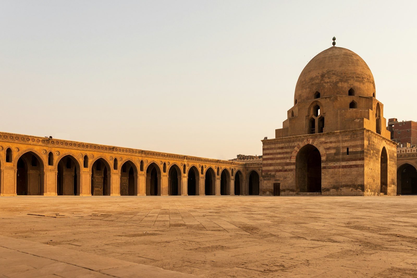 Patio vacío de la mezquita de Ibn Tulun, con una larga arcada de arcos apuntados y el edificio con cúpula de la fuente de abluciones, de muros a franjas, bajo una luz cálida.