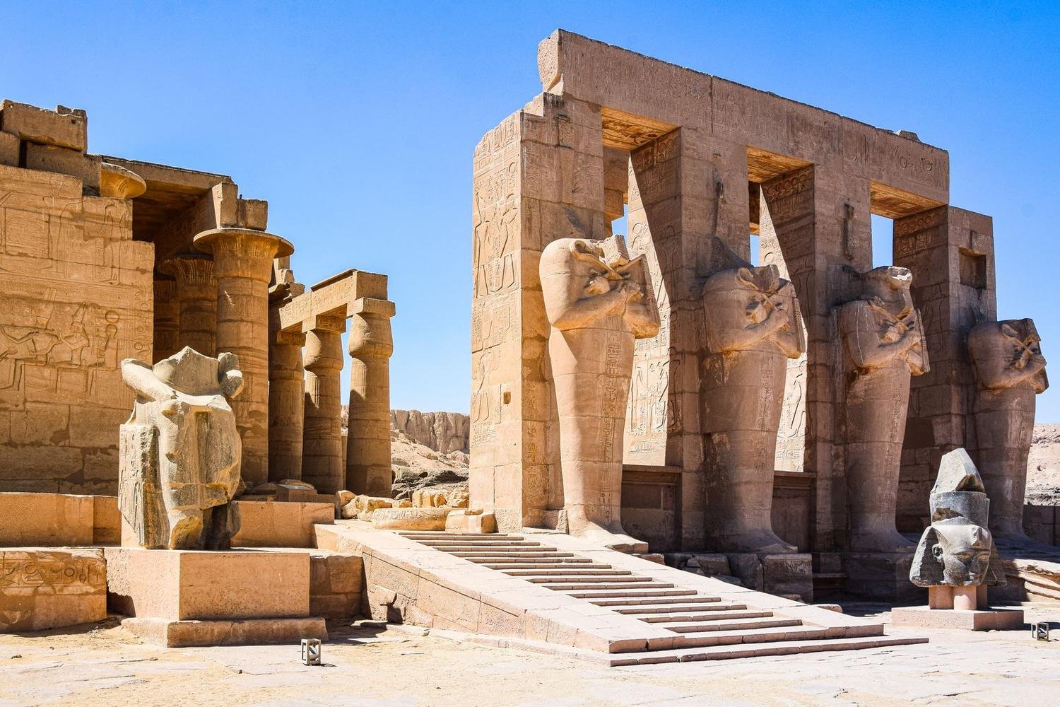 Patio de un templo de arenisca con estatuas osiríacas de brazos cruzados, muros cubiertos de jeroglíficos y una rampa de escalones bajo un cielo azul