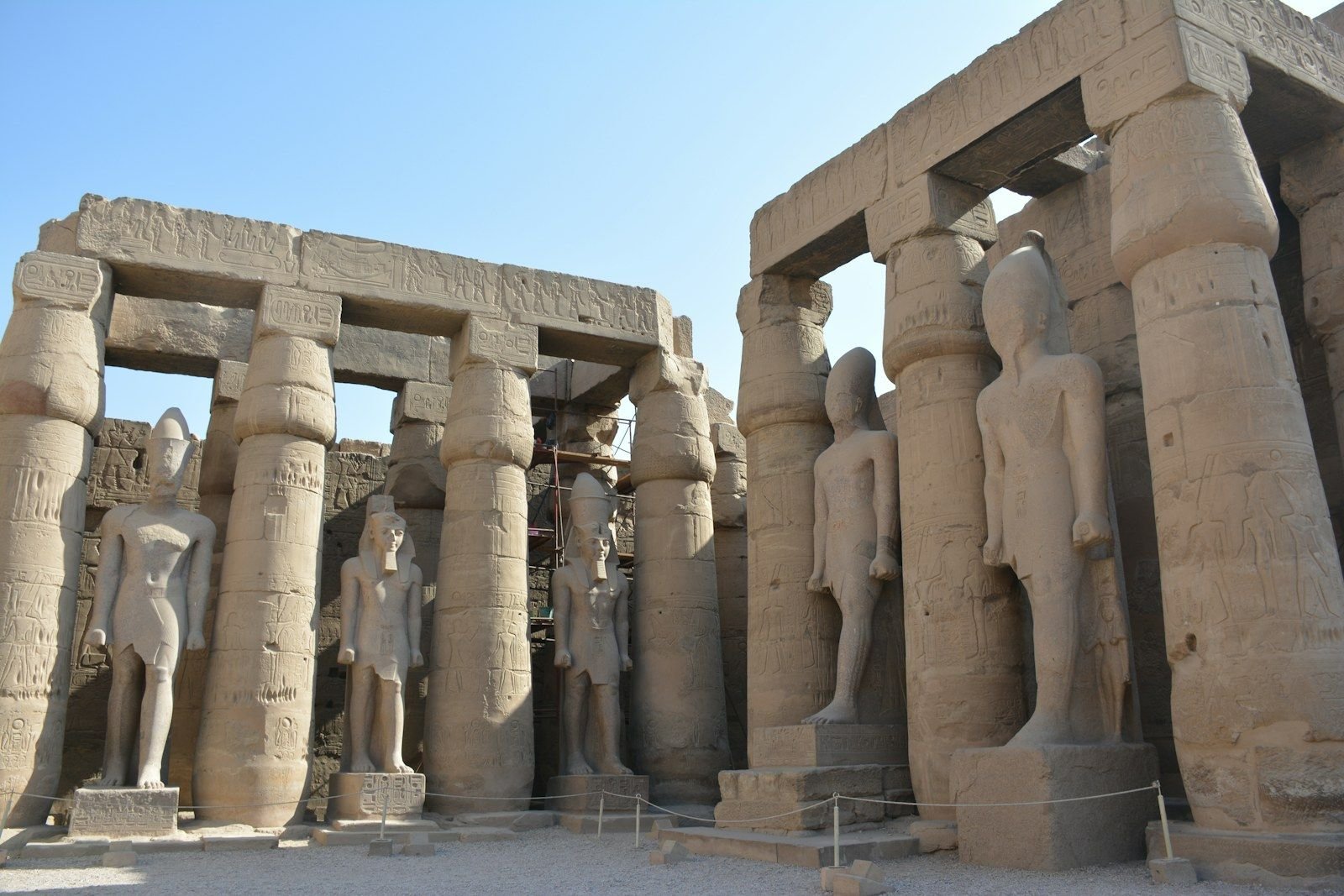 Estatuas de pie entre columnas con capiteles de papiro y vigas de piedra con relieves en el patio soleado de un templo antiguo