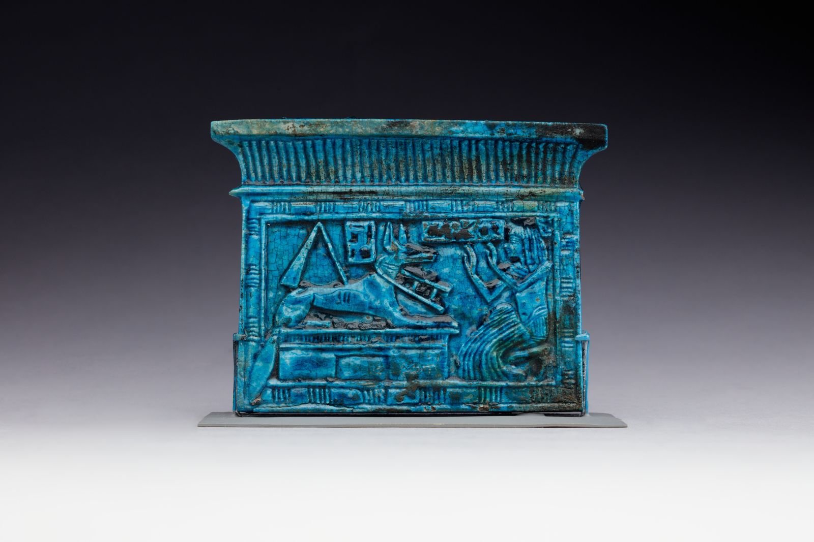Pectoral de loza vidriada azul con forma de capilla y un relieve de chacal tendido junto a una figura de rodillas con los brazos alzados