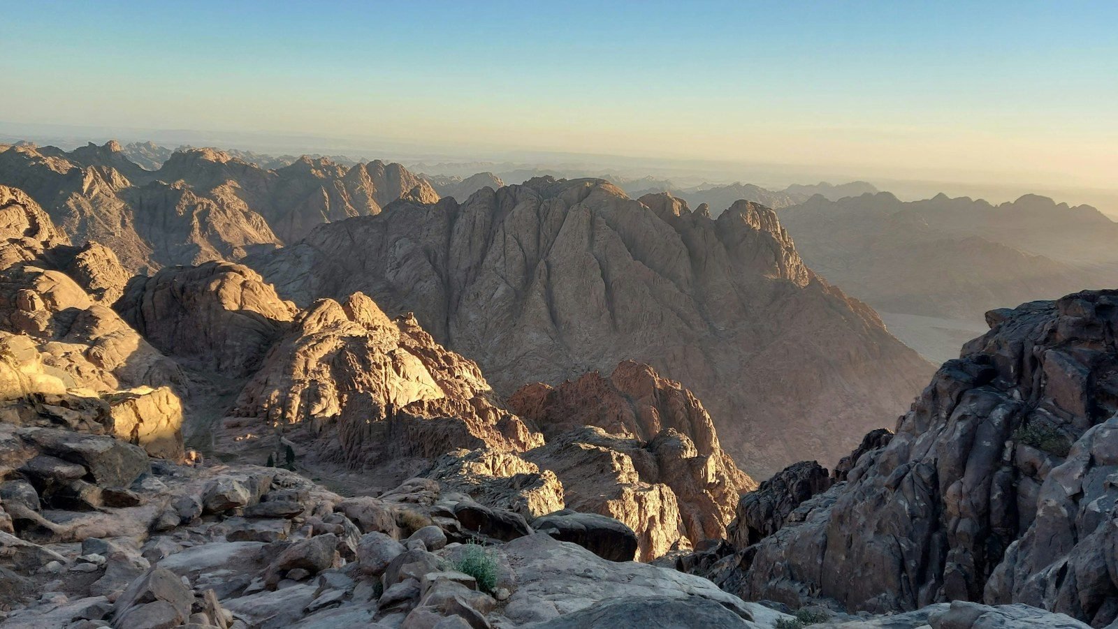 Picos de granito escarpados y un valle profundo bajo una luz cálida y baja, vistos desde una cornisa rocosa con cordilleras lejanas entre la bruma.