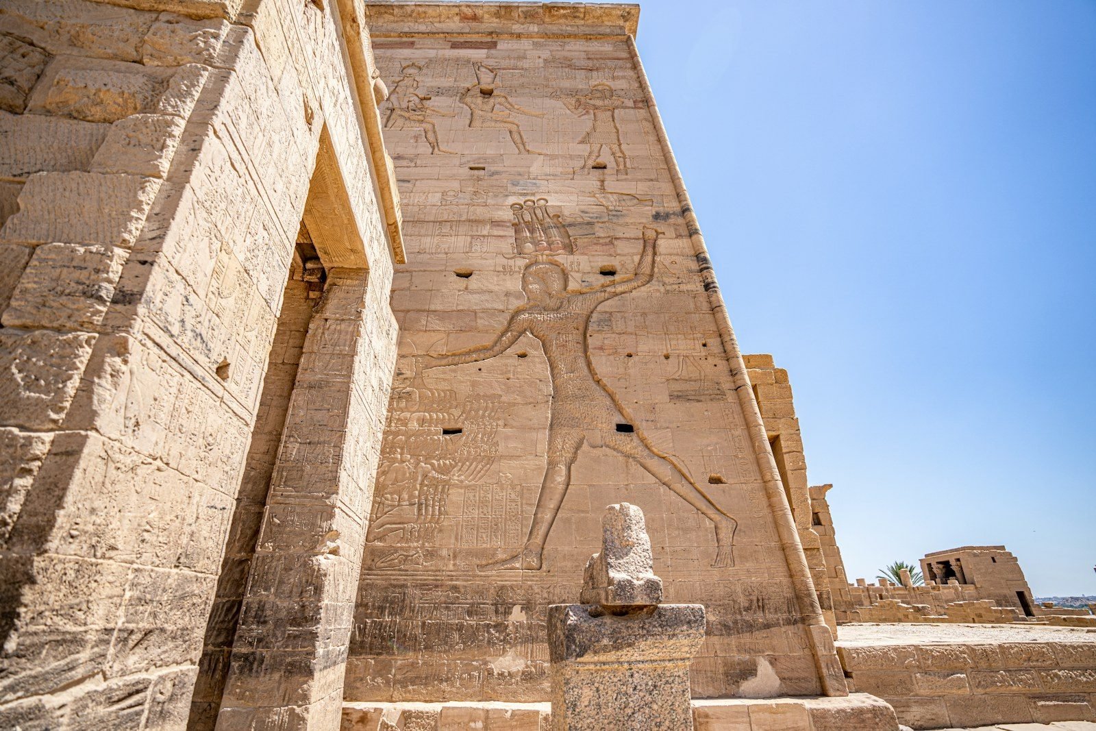 Torre de un pilono con un gigantesco relieve de un rey que golpea a sus enemigos, con una estatua de granito desgastada sobre un pedestal en primer plano.