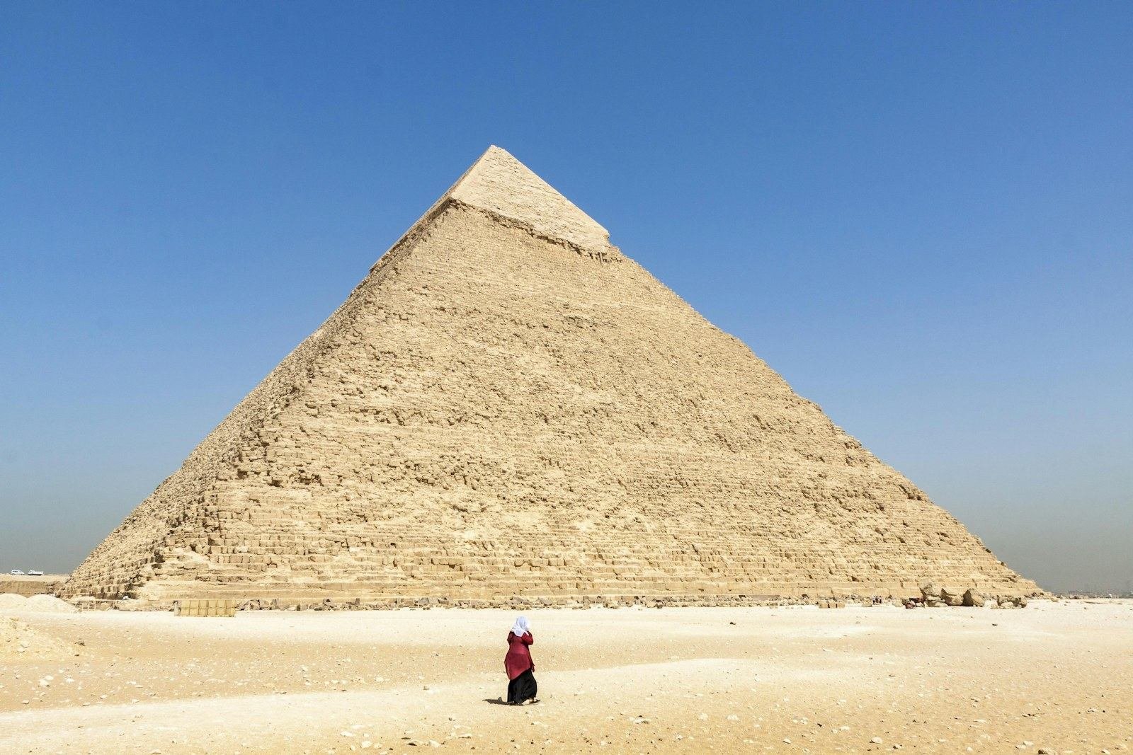 Una pirámide de piedra caliza con la cima lisa bajo un cielo azul despejado, y una persona que camina de espaldas por la arena clara del desierto