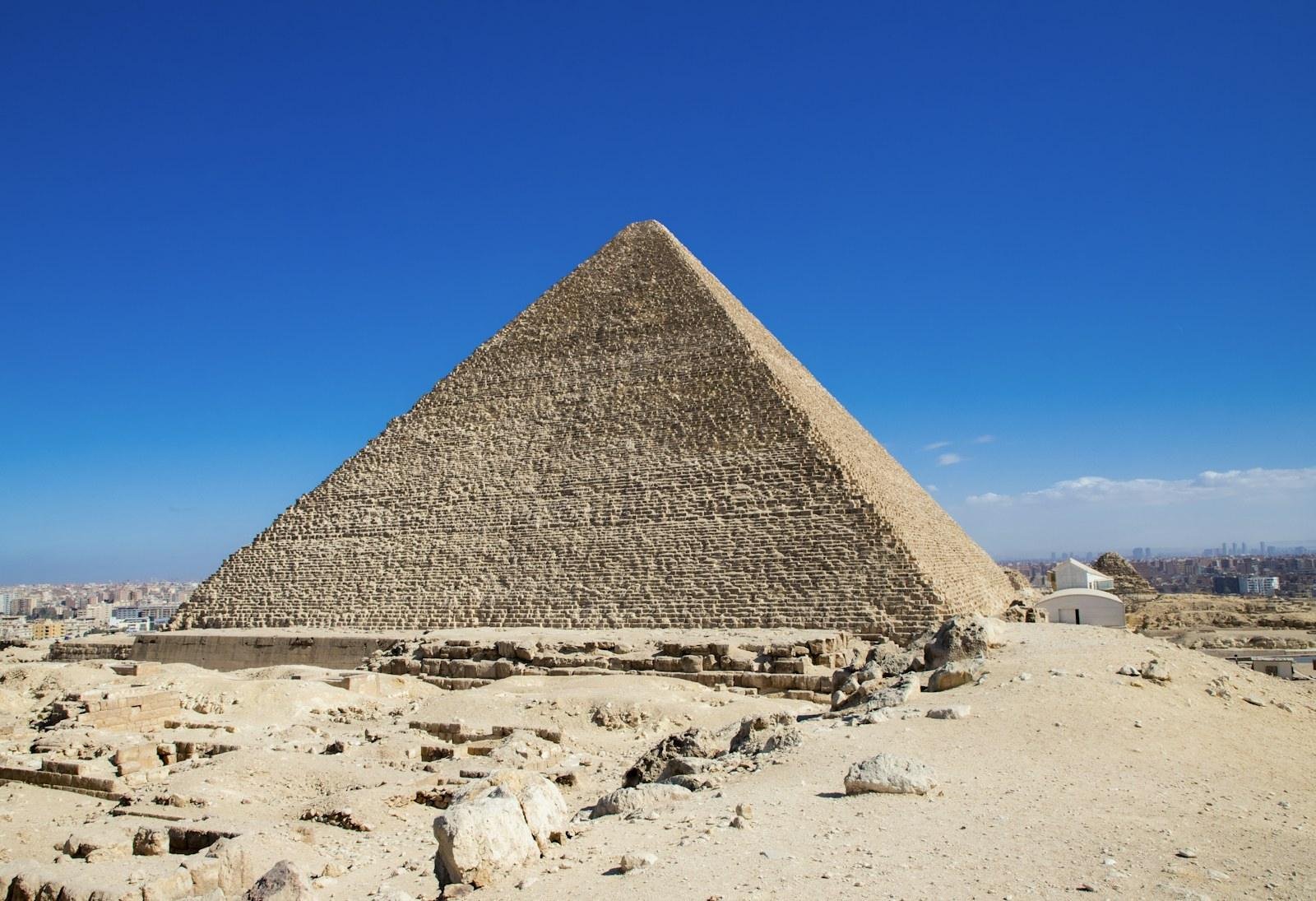 A large limestone pyramid on rocky ground with low ruined walls in front of it, and the city buildings far behind, under a deep blue sky