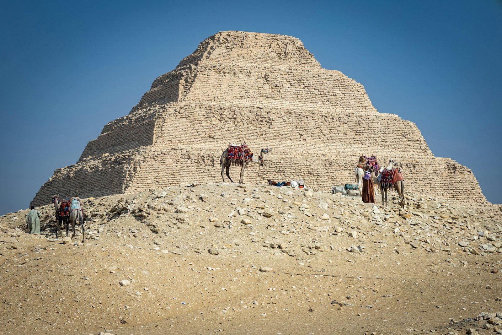 Una pirámide escalonada de piedra caliza detrás de un montículo de escombros, con tres camellos ensillados con mantas de colores y sus cuidadores al pie, bajo un cielo azul.