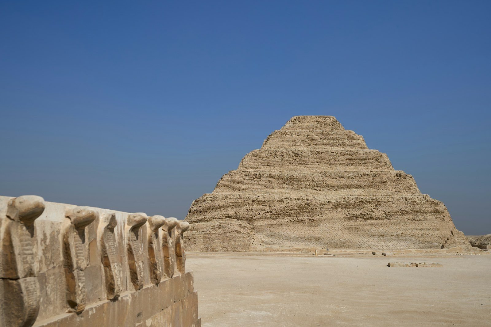 La pirámide escalonada de Djoser en Saqqara, con sus seis escalones, detrás de un muro rematado por una hilera de cobras erguidas talladas, bajo un cielo azul intenso.