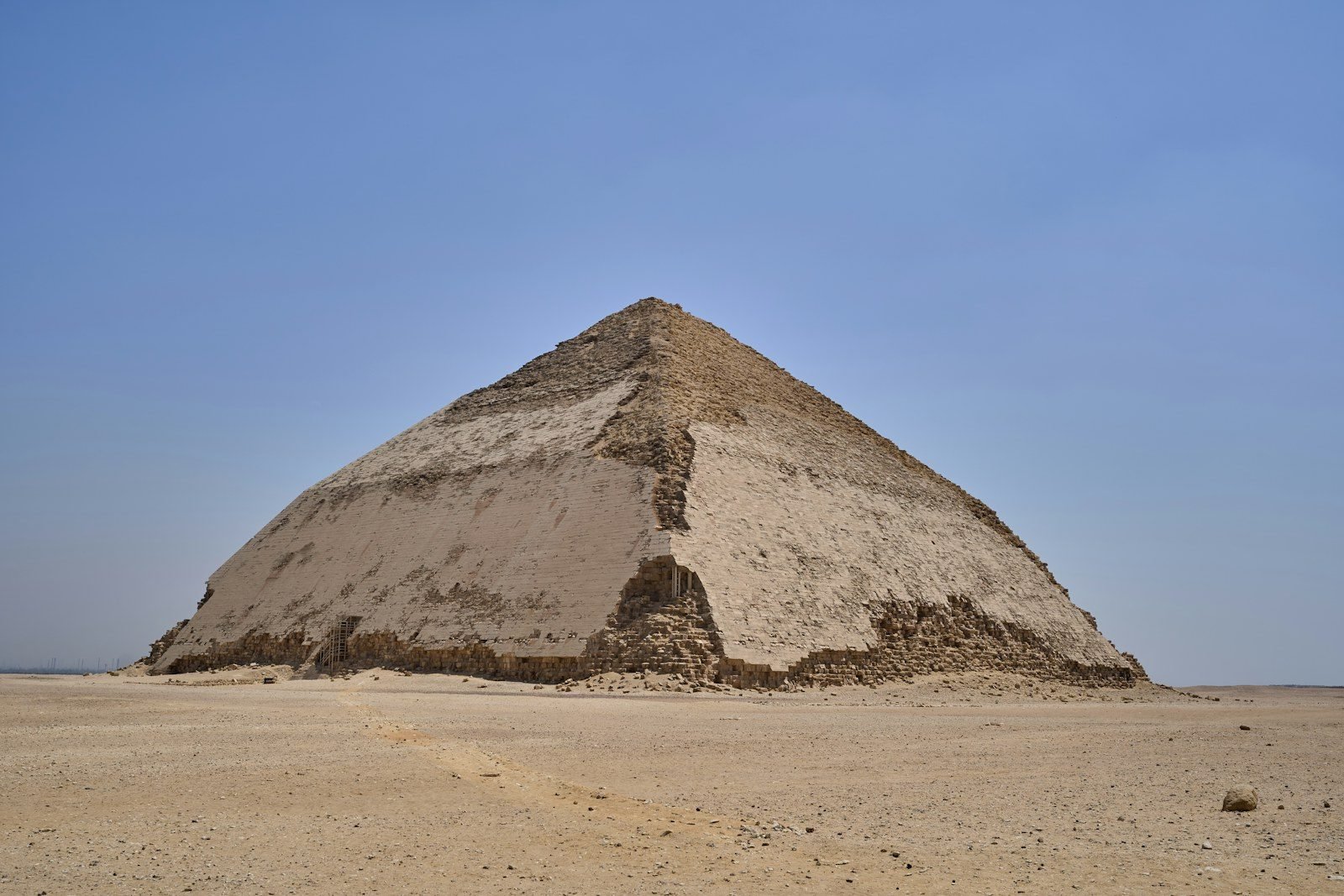 La Pirámide Romboidal de Dahshur vista desde una esquina, con el cambio de pendiente y zonas de revestimiento liso, sola sobre el desierto bajo un cielo azul con bruma.