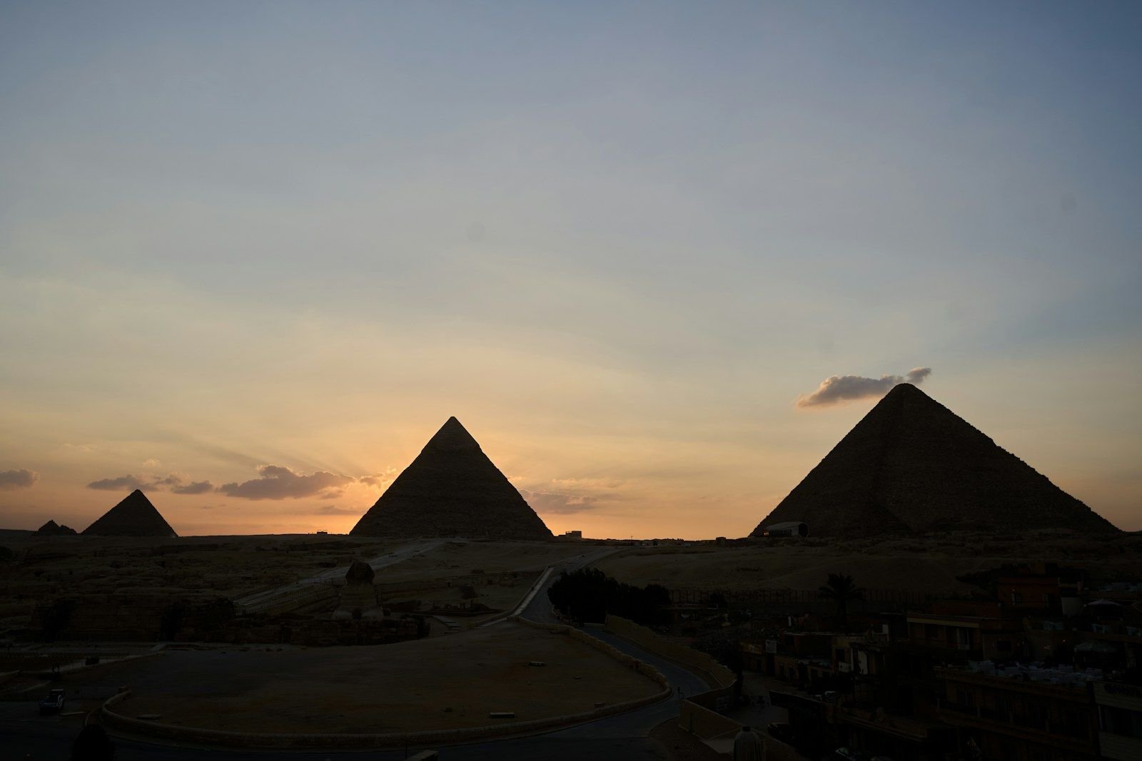 The pyramids of Giza at dusk as dark silhouettes against an orange and blue sky, with the buildings of the city in the foreground