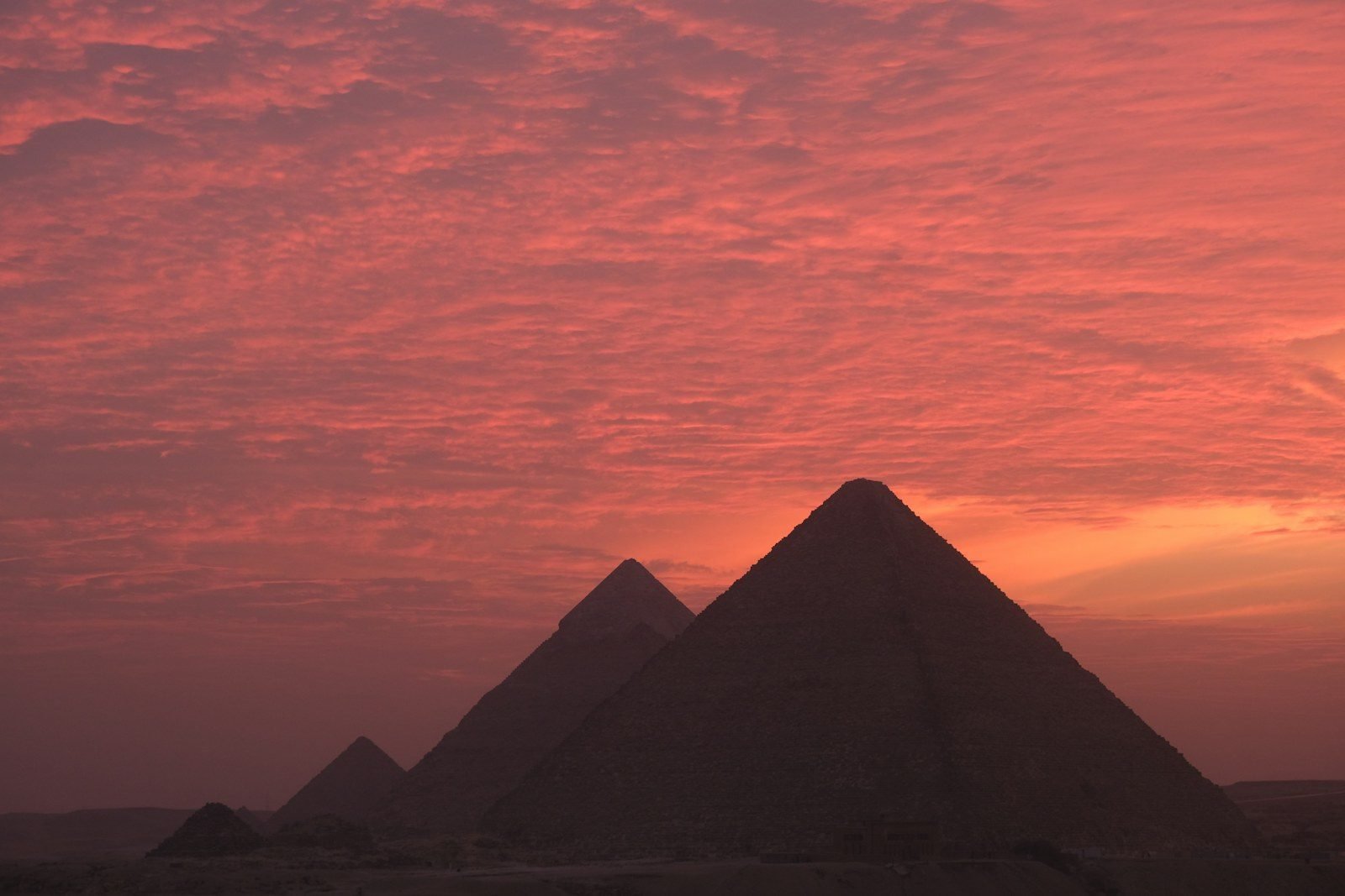 Las pirámides de Giza y las pirámides menores de las reinas, en silueta oscura contra un cielo de atardecer rojizo con nubes.