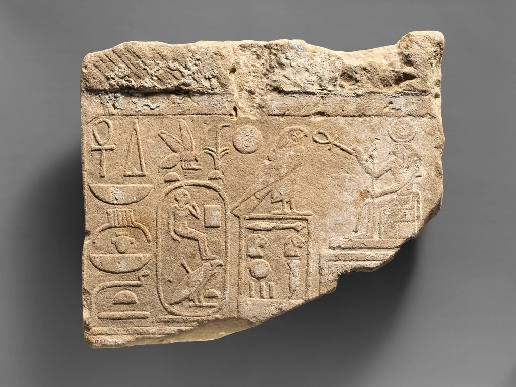 Fragmento de relieve de arenisca con jeroglíficos tallados, un cartucho real ovalado, un halcón sobre un recuadro y una figura sentada a la derecha.