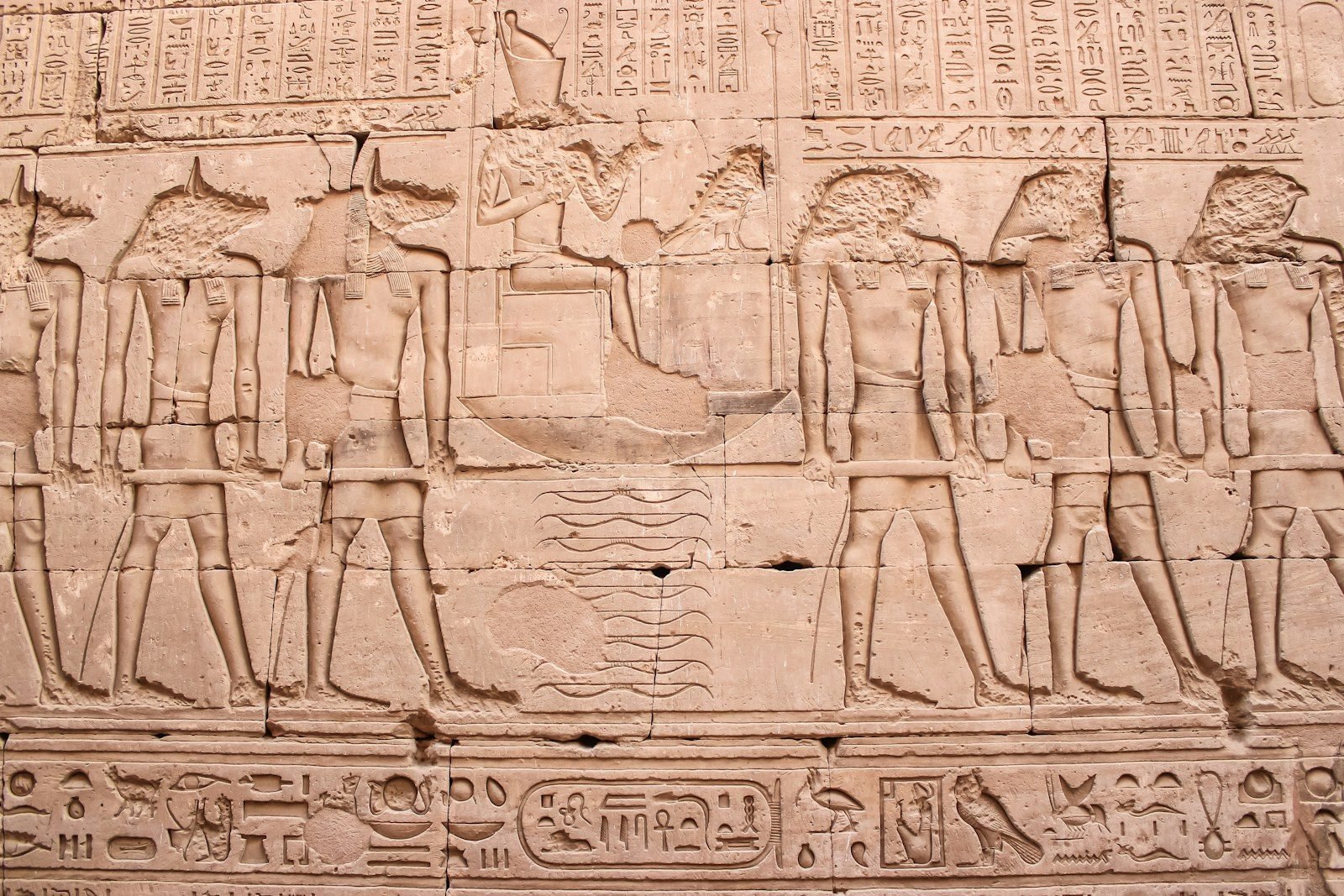 Relieve tallado en un muro de arenisca con figuras de cabeza de chacal y de halcón junto a una figura sentada en una barca, entre bandas de jeroglíficos.