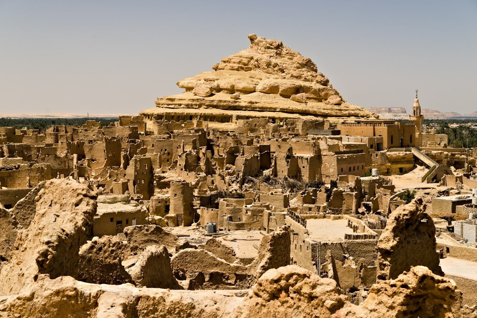 Ruinas de adobe de una antigua ciudad de oasis a los pies de una colina de roca estratificada, con una mezquita y su minarete a la derecha.