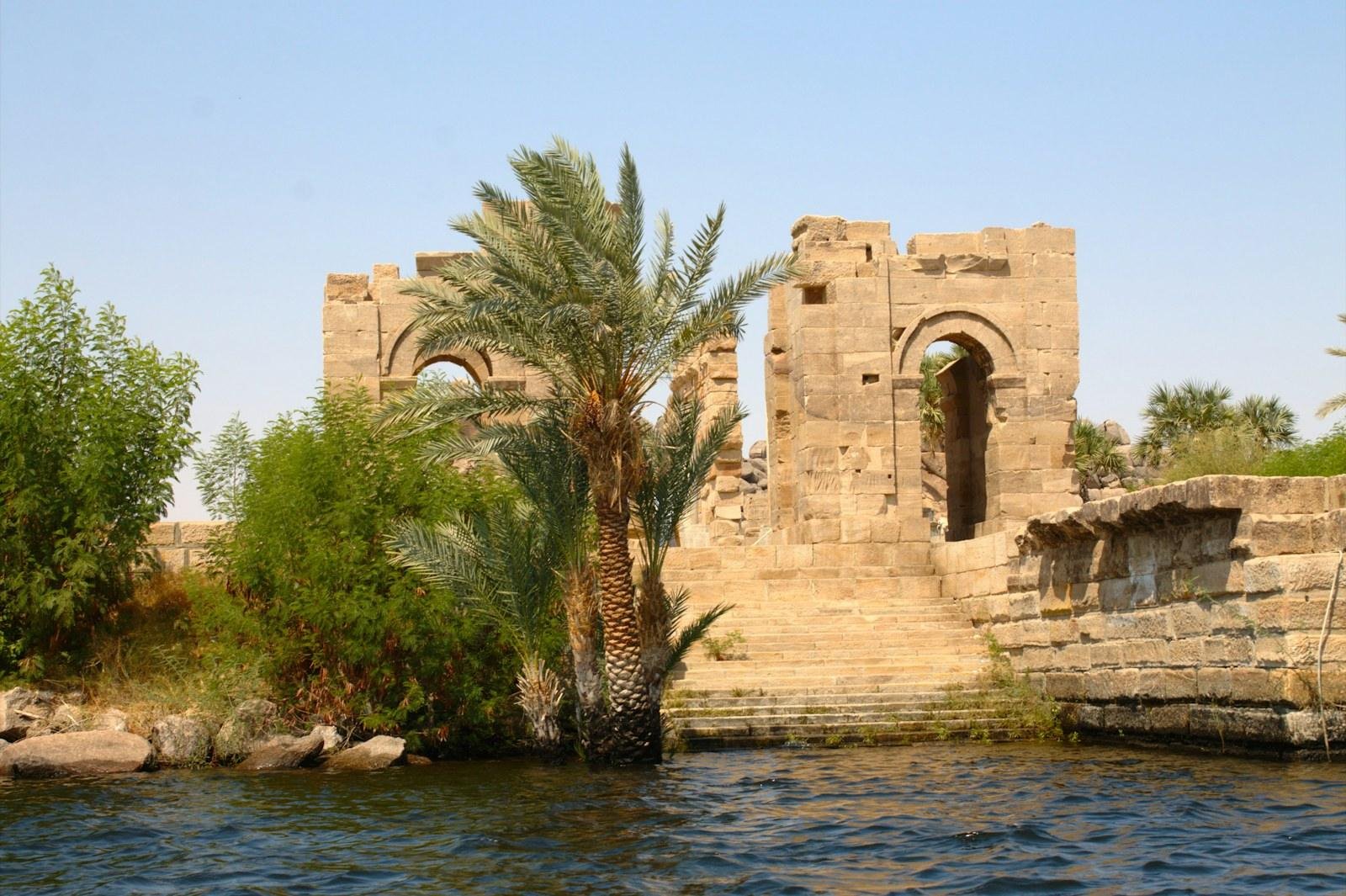 Weathered sandstone ruins with arched doorways and a stone stairway reach the water's edge beside tall date palms and green bushes