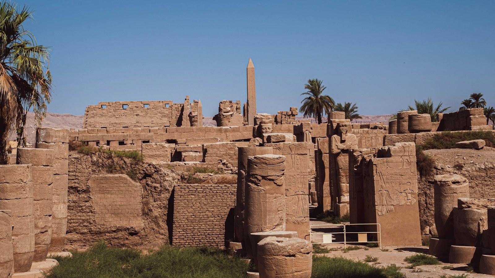 Muros de un templo de arenisca con un obelisco y palmeras bajo un cielo azul despejado, como el de Luxor en verano