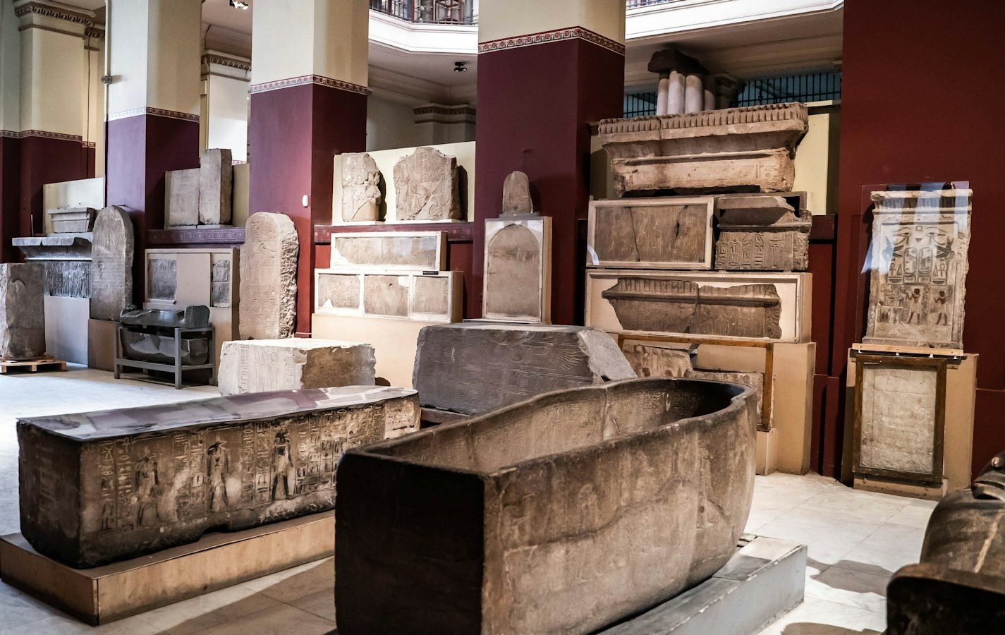 Sala de un museo con sarcófagos de piedra, estelas y bloques con relieves expuestos entre columnas granates sobre un suelo de mármol blanco, sin visitantes.