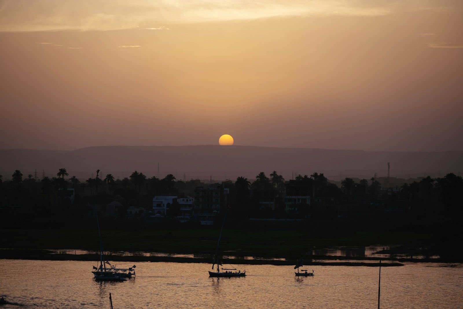 El sol bajo y anaranjado se pone sobre una orilla de palmeras entre la bruma, mientras pequeños veleros descansan en el agua del río