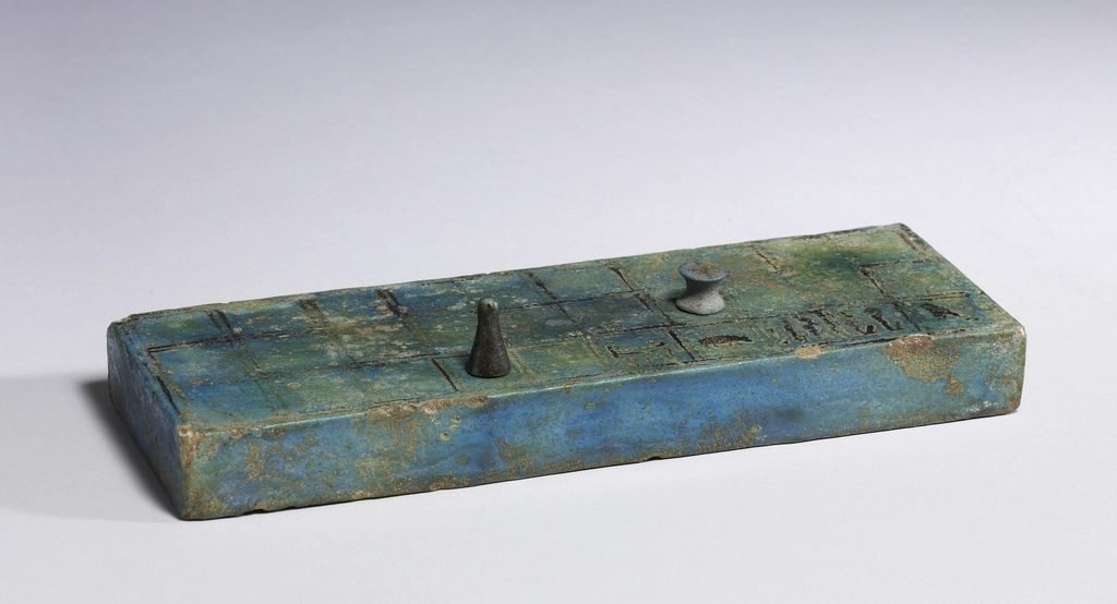 Tablero de senet esmaltado en azul verdoso con dos fichas encima, sobre un fondo gris claro.
