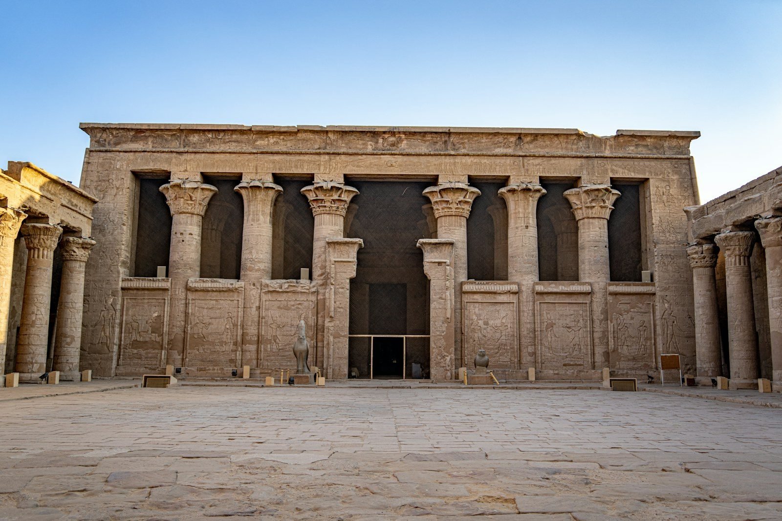 Fachada del templo de Horus en Edfú con sus columnas de capiteles florales, muros de relieves y dos estatuas de halcón a la entrada, vista desde el patio.