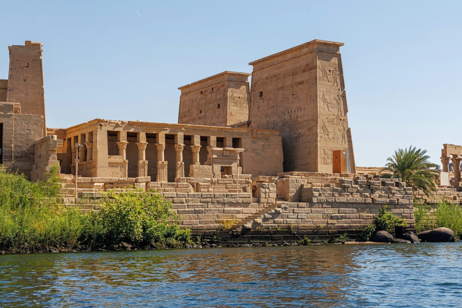El pilono y la columnata del templo de Isis en Filae vistos desde el agua, sobre un basamento de bloques de piedra con arbustos y una palmera.