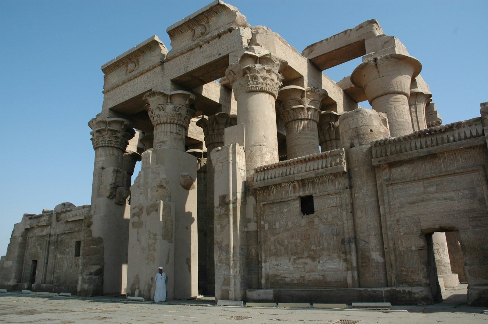 Floral-capital columns and the cornice of the double temple of Kom Ombo seen from the courtyard under a blue sky, with one small figure beside a pillar.