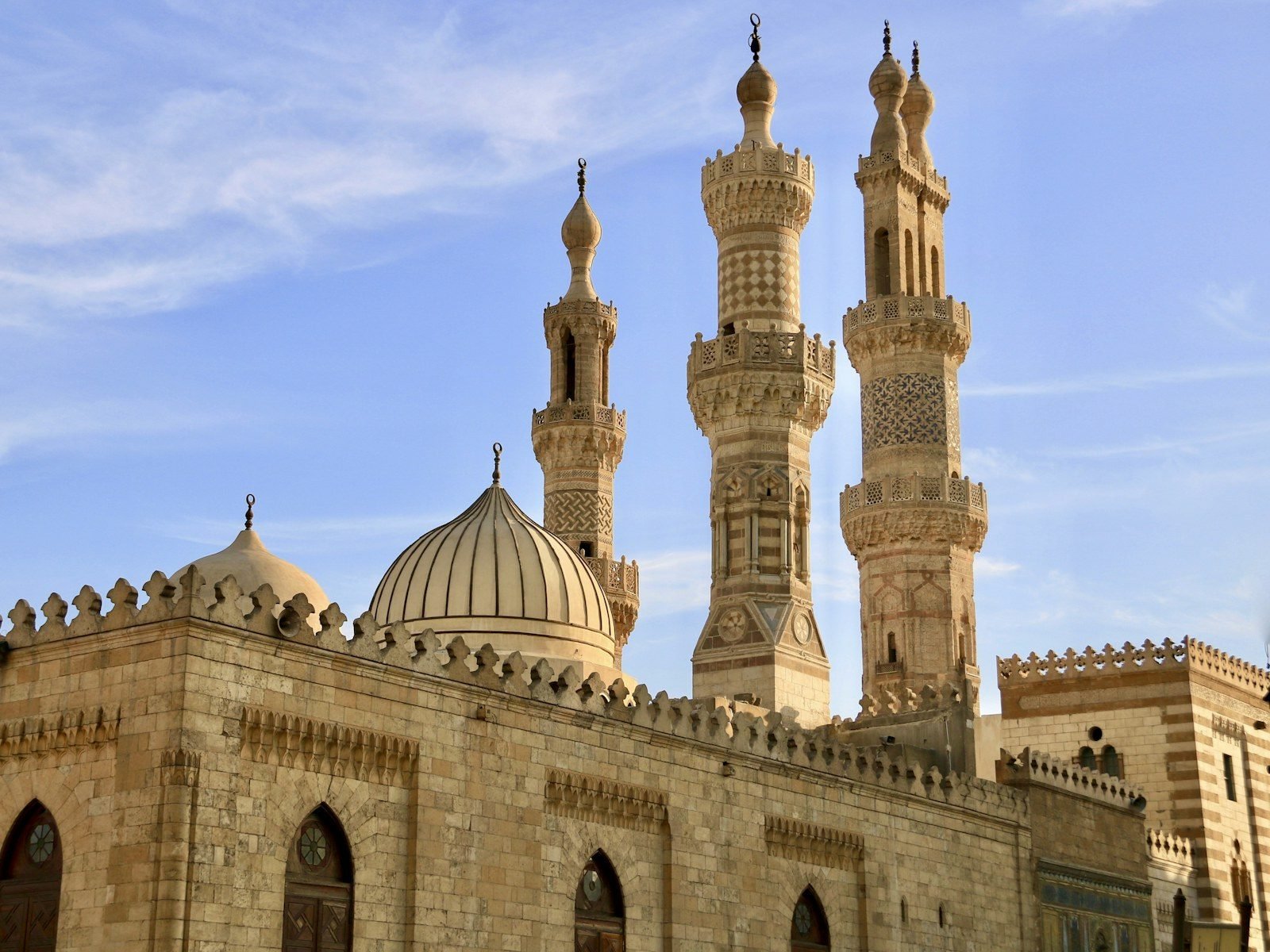 Tres minaretes de piedra tallada con fustes decorados y una cúpula acanalada se alzan tras un muro almenado bajo un cielo azul con nubes finas.