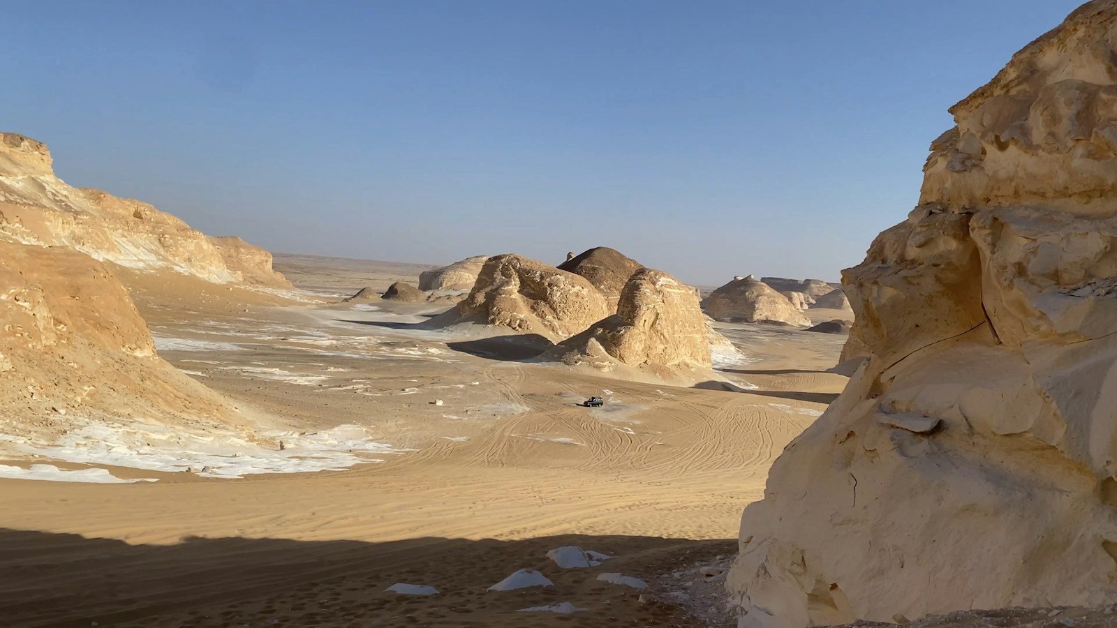 Valle desértico con formaciones de roca blanca y arenisca dorada enmarcadas por un gran bloque claro, y un pequeño todoterreno sobre la arena marcada por huellas.
