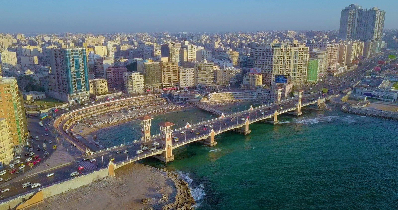 Aerial view in golden light of a curved bridge with two ornate towers crossing a small bay lined with beach cabins, beside the apartment blocks of a large coastal city.